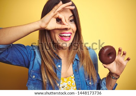 Young beautiful woman eating chocolate donut over yellow background with happy face smiling doing ok sign with hand on eye looking through fingers