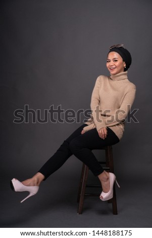 Portrait of a beautiful Muslim female model wearing sweatshirt and jeans with hijab sitting on a chair isolated over grey background. Studio fashion and beauty concept.