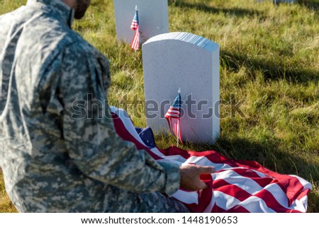 selective focus of soldier in camouflage uniform holding american flag and sitting in graveyard 