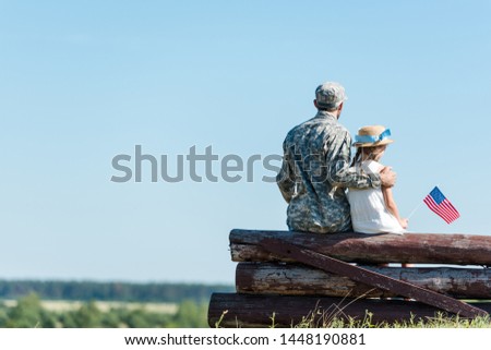 back view of patriotic child holding american flag near veteran father while sitting in fence 