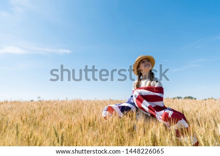 smiling kid in straw hat holding american flag in golden field with wheat 