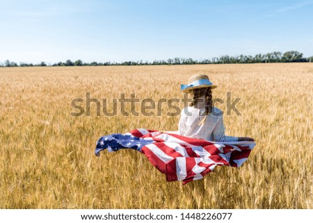 back view of kid in straw hat holding american flag in golden field in summertime 