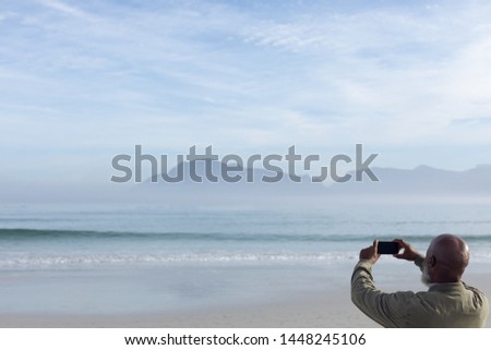 Rear view of handsome senior African-American man taking a picture of the sea and mountains on beach on beautiful day. Authentic Senior Retired Life Concept