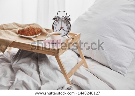 alarm clock, sticky note with happy morning lettering, coffee and croissant on wooden tray on white bedding