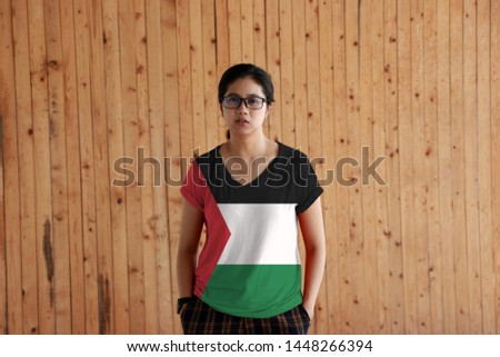 Woman wearing Palestine flag color shirt and standing with two hands in pant pockets on the wooden wall background, black white and green; with a red triangle based at the hoist. 