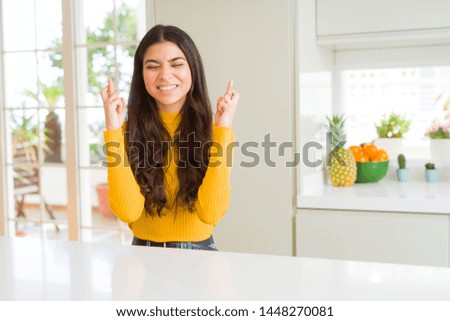 Young beautiful woman at home on white table smiling crossing fingers with hope and eyes closed. Luck and superstitious concept.