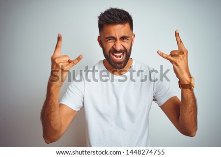 Young indian man wearing t-shirt standing over isolated white background shouting with crazy expression doing rock symbol with hands up. Music star. Heavy music concept.