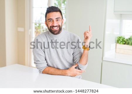 Handsome hispanic man wearing casual sweater at home with a big smile on face, pointing with hand and finger to the side looking at the camera.