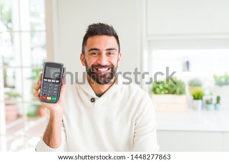 Handsome hispanic man holding point of sale terminal dataphone with a happy face standing and smiling with a confident smile showing teeth