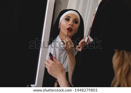 Woman in a nuns suit paints lips in red near a mirror