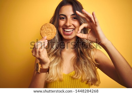 Young beautiful woman eating biscuit over grey isolated background doing ok sign with fingers, excellent symbol