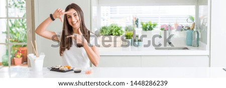 Wide angle picture of beautiful young woman eating asian sushi from delivery smiling making frame with hands and fingers with happy face. Creativity and photography concept.