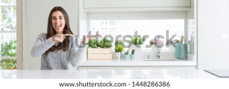 Wide angle picture of beautiful young woman sitting on white table at home amazed and smiling to the camera while presenting with hand and pointing with finger.