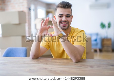 Young man sitting on the table with cardboard boxes behind him moving to new home smiling in love showing heart symbol and shape with hands. Romantic concept.