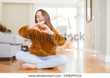 Beautiful young girl kid sitting on the floor at home smiling in love showing heart symbol and shape with hands. Romantic concept.