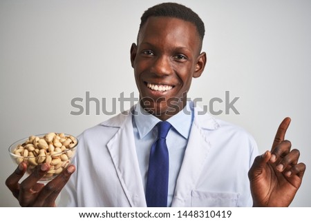 African american doctor man holding bowl with pistachios over isolated white background very happy pointing with hand and finger to the side