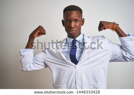 Young african american doctor man wearing coat standing over isolated white background showing arms muscles smiling proud. Fitness concept.