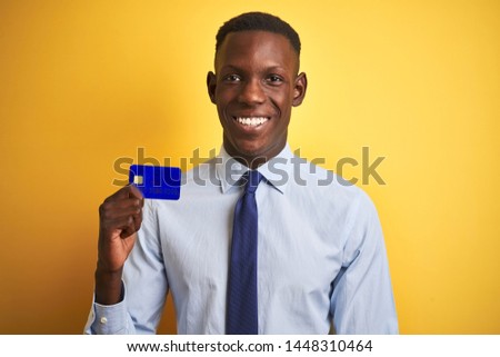 African american businessman holding credit card standing over isolated yellow background with a happy face standing and smiling with a confident smile showing teeth