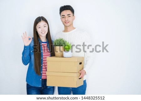Beautiful young asian couple over white background holding cardboard boxes doing ok sign with fingers, excellent symbol
