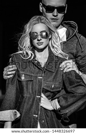 Stilish couple cuddling in modern blue jeans jackets and trendy sunglasses on summer city background. Beautiful female blond model. Closeup portrait. Black and white