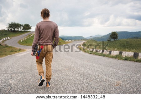 A stylish young man walks along a winding mountain road with a skate or longboard in his hands the evening after sunset. The concept of youth sports and travel hobbies