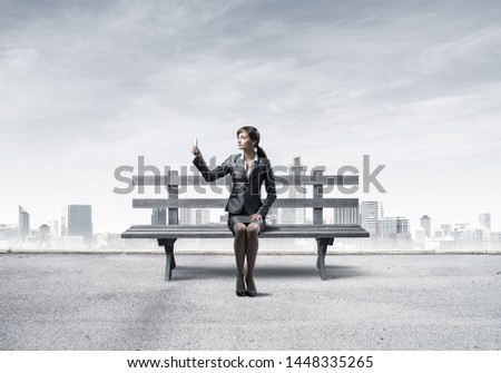 Business woman taking selfie photo or chatting with smartphone. Attractive girl using mobile phone on wooden bench. Mobile marketing and communication. Modern cityline panorama with cloudy sky.