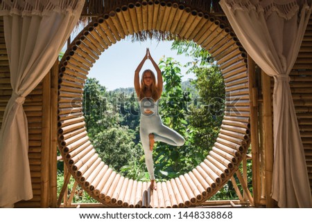 Woman practising yoga (tree pose) in bamboo house studio. View of nature and mountains on background. Peaceful meditation spot. Ecolodge retreat, eco friendly house Royalty-Free Stock Photo #1448338868