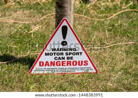 Red triangle warning sign alerting spectators to the risks of motor sports. Black and red text, DANGEROUS in bold with large exclamation mark. The signs leans against a post set in the grass.
