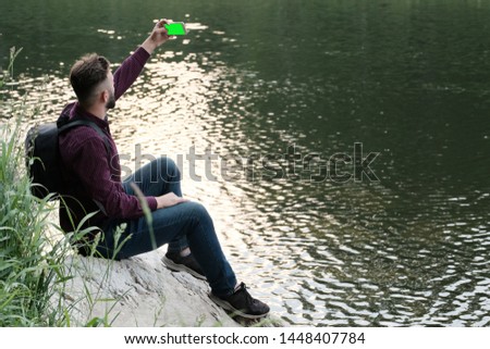 A guy with a phone in his hands sits on a stone by the river. Traveler with a backpack and a smartphone makes selfie or shooting online. Concept of travel, tourism and lifestyle. Copy space.