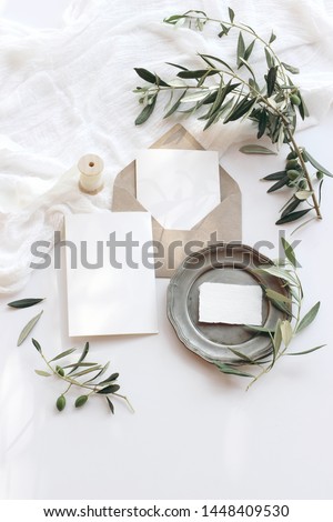 Summer wedding stationery mock-up scene. Blank greeting cards, envelope, vintage silver plate, olive branches and ribbon. White background with cotton table runner in sunlight. Flat lay, top view.