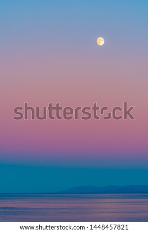Moon with pink skies at blue hour twilight before sunrise/ following sunset over ocean with reflection in water and mountain silhouette in background. Colorful scenic nature and landscape setting.