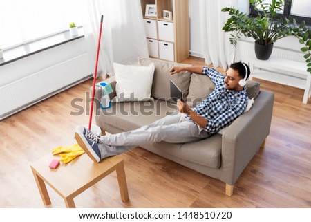 people, housework and housekeeping concept - indian man in headphones listening to music on tablet computer after home cleaning