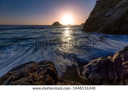 The sunburst with Seal Rock in the foreground at Pt Lobos, San Francisco, California with waves rushing in to the rocks on the beach