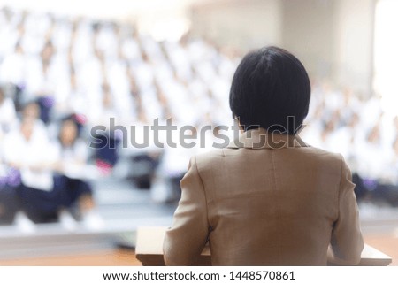 The rear view of the female teacher is do the presentation behind the podium with blurred audience background.