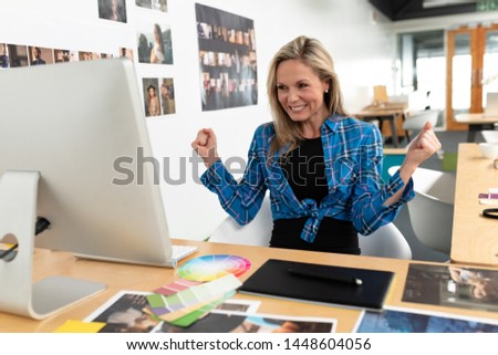 Front view of happy Caucasian female graphic designer celebrating success at desk in office. This is a casual creative start-up business office for a diverse team