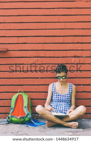 Barefoot girl draws sitting near a brick wall. Brunette against the background of a red wall. A woman with a backpack walks along a city street. The artist makes a sketch.