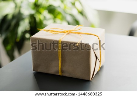 Office snacks delivery service. Closeup of beige paper gift box tied with yellow cord. Blur green plant in background. Copy space.