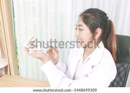 Smiling pretty Asian woman dentist setting in office clinic, holding dental equipment and considering teeth model on table. Concept of female professional in medical healthcare career. 