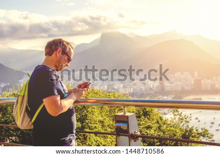 A guy with a backpack takes picture on mobile phone on the viewpoint at Urka and Sugar Loaf. View of Rio de Janeiro, Brazil. Tourist man at sunset.