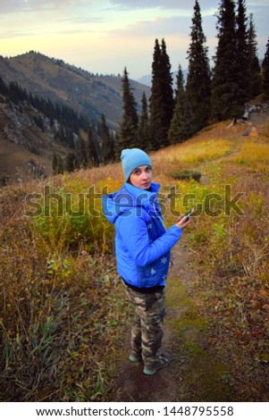 High angle view of a girl in the mountain forest with a mobile phone in hand. She looks surprised or frightened or even lost. Pretty girl in warm clothes lost in the mountains in the evening.