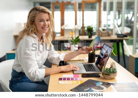 Portrait of Caucasian female graphic designer holding a color swatch while using laptop at desk in office. This is a casual creative start-up business office for a diverse team