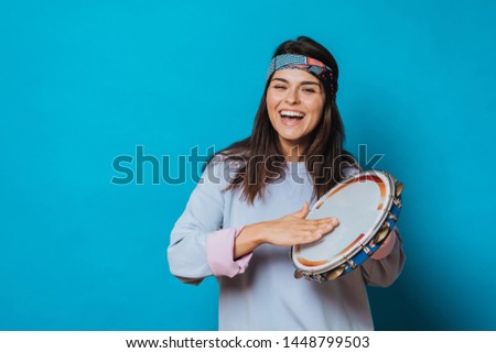 Cheerful girl with a headband on her head and grey sweater playing on tambourine and laughing happily over blue background. People isolated. Royalty-Free Stock Photo #1448799503