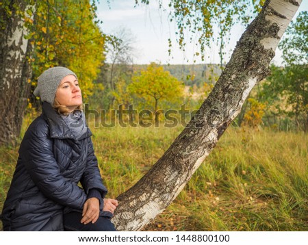 A pensive girl sitting on a birch tree on the outskirts of a withering autumn forest.