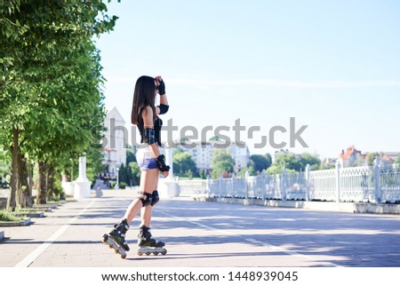 Young brunette woman with long hair, wearing rollerblades, posing in city park in the morning. Standing full-length portrait of roller-skater, wearing black top and white shorts. Summer activities. 