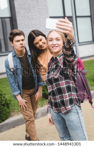 positive teenagers holding smartphone, taking selfie and smiling outside 