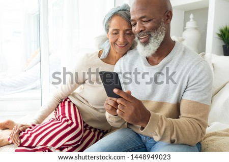 Front view of happy diverse senior couple using a smartphone on sofa in beach house. Authentic Senior Retired Life Concept Royalty-Free Stock Photo #1448949023