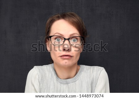 Studio close-up portrait of cute and funny caucasian fair-haired girl with eyeglasses, silly grimacing and looking naively and with bewilderment at camera, against gray background. Headshot Royalty-Free Stock Photo #1448973074