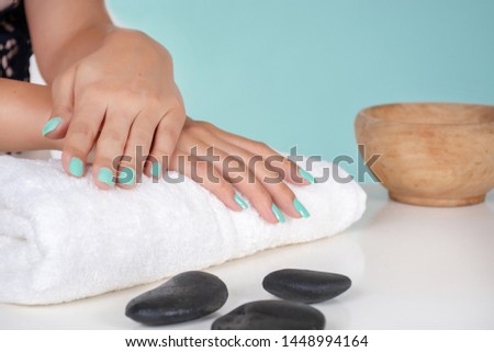Young girl hands with a turquoise color nails polish on a towel and decorative stone on desk isolated on soft blue background in studio. Manicure and beauty concept. Close up, selective focus