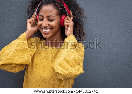 African girl listening to music with her wireless headphones while leaning against grey wall. Happy beautiful young woman listening music with closed eyes enjoying the melody on gray background.