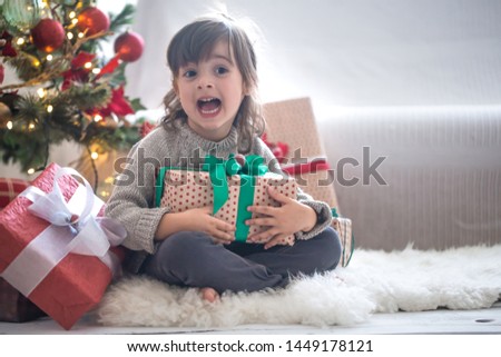 Pretty little girl is holding a gift box and smiling while sitting on her bed in her room at home. Happy Holidays. Gifts and presents